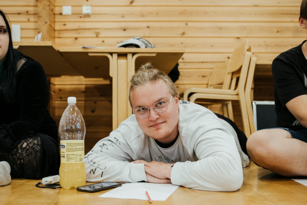 Kolme nuorta, joista kaksi istuu ja keskimmäinen loikoilee salin lattialla käsivarsiinsa nojaten.