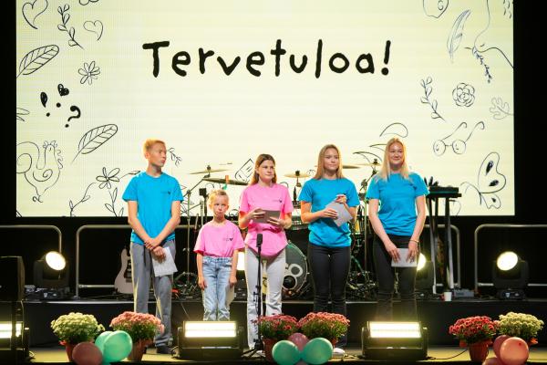 suomeksi: Eri-ikäisiä oppilaita lavalla, taustalla Tervetuloa-kyltti. på svenska: Elever i olika åldrar står på en scen, med en skylt med texten 