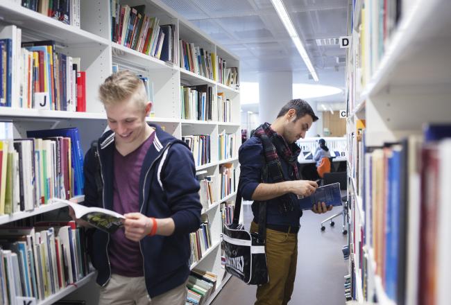 Kaksi miestä selailee kirjoja kirjaston hyllyjen välissä