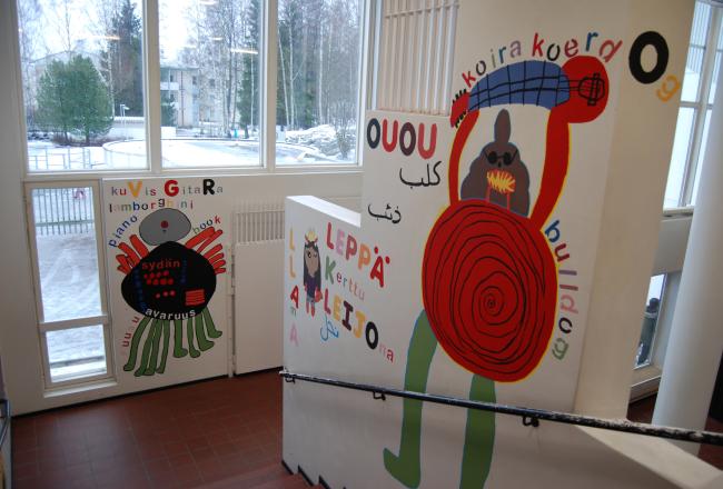 Oppilaiden koulun seinille maalaamia hahmoja joiden ympärille on kirjoitettu sanoja koulussa käytetyillä eri kielillä