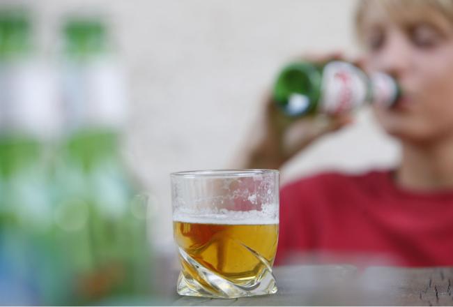  I förgrunden på bilden står ett glas öl, i bakgrunden dricker en ung öl ur en flaska.