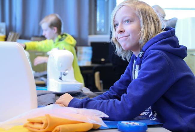 Poika hymyilee ompelukoneen äärellä. Pojalla on vaaleat pitkät hiukset ja sininen huppari.