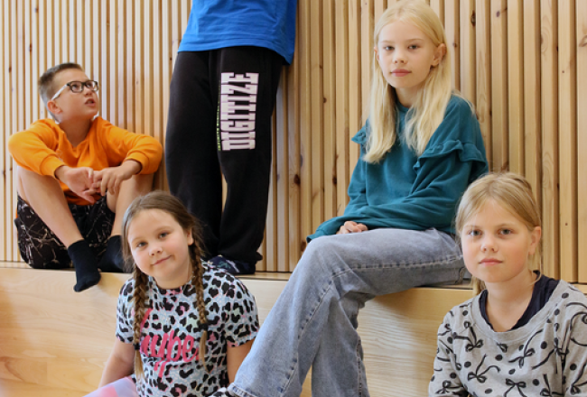 Kaksi lasta istuu penkillä ja heidän välissään yksi lapsi seisoo penkillä. Lasten edessä lattialla istuu kaksi lasta.