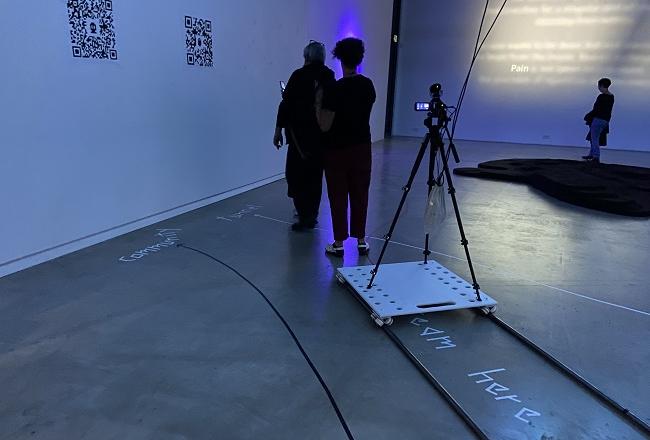 Gallerian sinertävässä valossa ihmisiä, tilaan kirjoitettuja ja heijastettuja tekstejä, QR-koodeja ja teknisiä laitteita