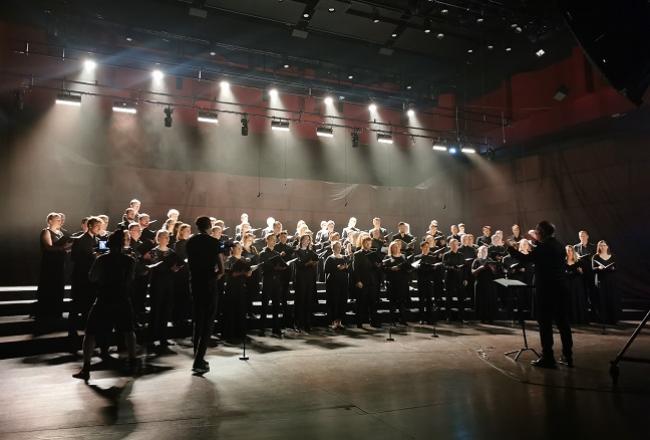 Kuoro esiintyy dramaattisissa valoissa takanaan tumma tausta