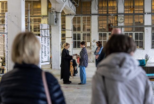 Tapahtuma vanhassa konepajahallissa. Etualalla kaksi osallistujaa selin, kuvan keskellä kaksi henkilöä keskustelevat vastatusten. Taustalla muita osallistujia sekä mustavalkoista tekstiä sisältäviä tekstiplansseja.