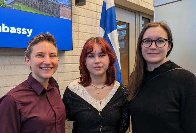 Kolme henkilöä seisoo Suomen lipun ja "embassy"-kyltin edessä.