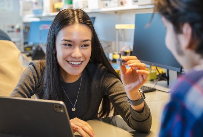 Nainen hymyilee ja pitää kädessään pientä teknistä laitetta. Hän keskustelee toisen henkilön kanssa tietokoneen äärellä, ja taustalla on tekninen laboratorioympäristö.