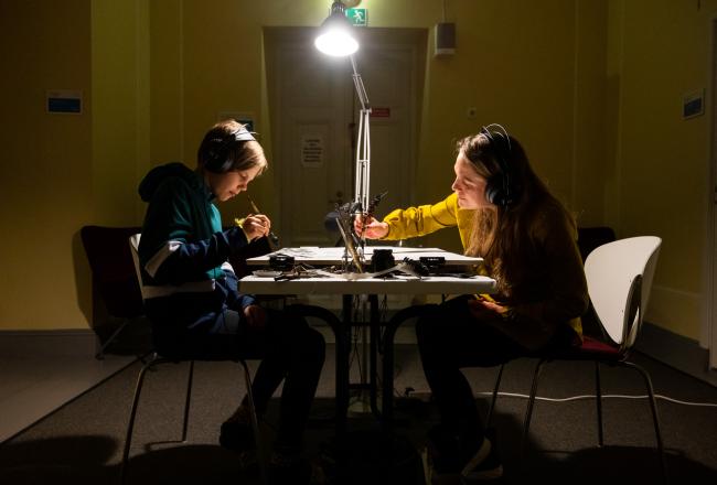 Kuva lapsesta ja aikuisesta naisesta pöydän ääressä kuuntelemassa musiikkia kuulokkeilla ja keskittyineinä kirjoittamiseen.