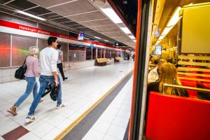 Kävelijöitä metroasemalla ja metron ikkunan läpi näkyviä matkustajia.