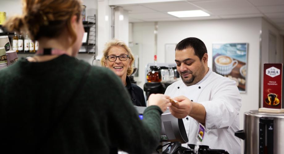 Asiakas, opettaja ja opiskelija ammatillisen oppilaitoksen herkkukaupan kassalla.