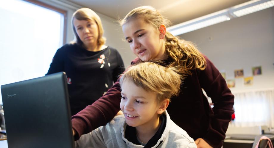 Poika ja tyttö opiskelemassa tietokoneella. Opettaja katselee taustalla.