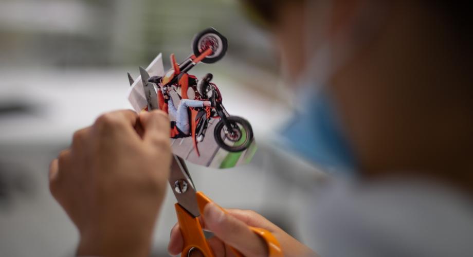 Kuvan keskellä näkyvät sakset, joilla opiskelija parhaillaan leikkaa irti motocross-ajajaa tulostepaperista. Muu kuvasta on epäterävä.