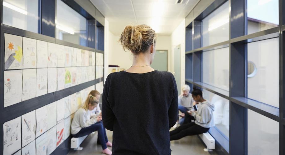 Nainen seisoo käytävällä selin. Taustalla näkyy lapsia kännykät kädessä.