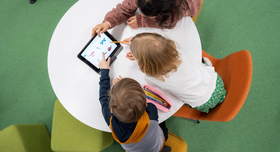 Kolme lasta istuu pöydän äärellä ja katsoo tablettia.
