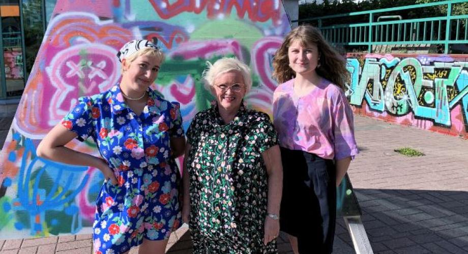 Kaksi nuorta ja vanhempi henkilö seisovat graffitiseinän edessä.