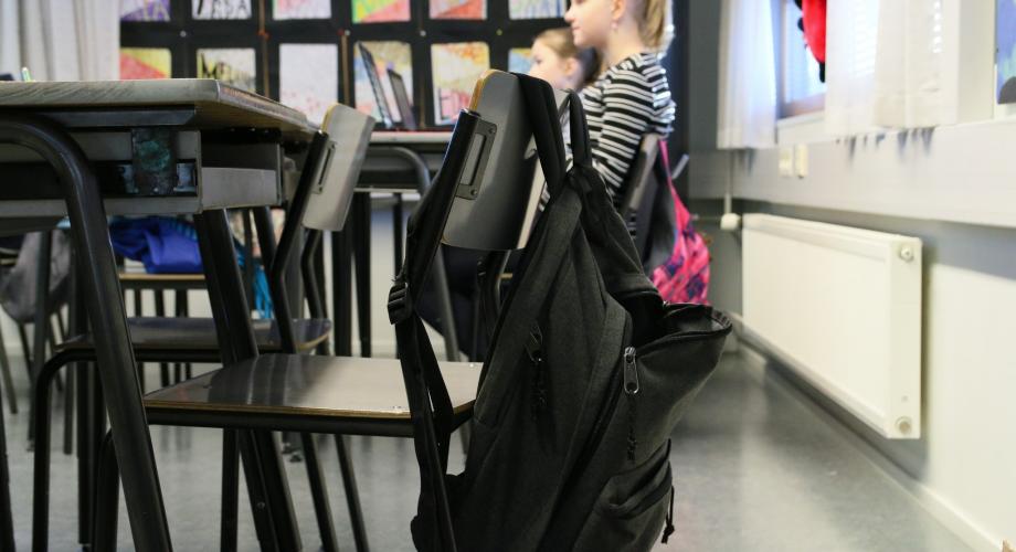 Luokkahuone, jossa etualalla pulpetti ja tyhjä tuoli, jonka selkänojalla roikkuu reppu. Tyhjän pulpetin takana on toinen pulpetti, jonka ääressä istuu kaksi tyttöä.