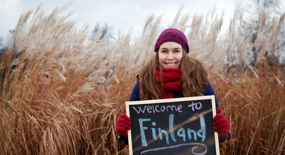 En kvinna med en mössa håller en skylt i ett höfält. På skylten står det Welcome to Finland.