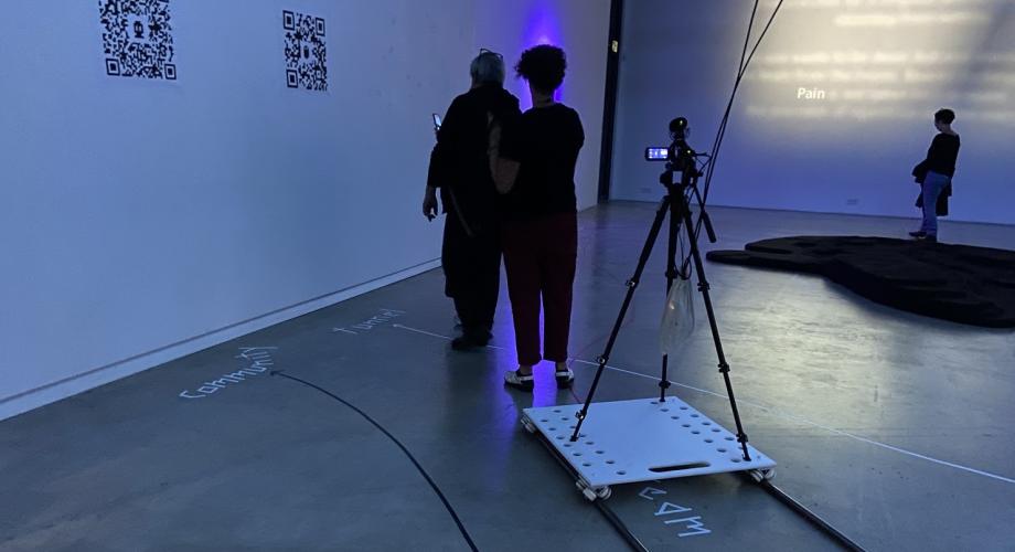 Gallerian sinertevässä valossa ihmisiä, seinään ja lattiaan heijastettuja ja kirjoitettuja tekstejä, QR-koodeja sekä erilaisia laitteita