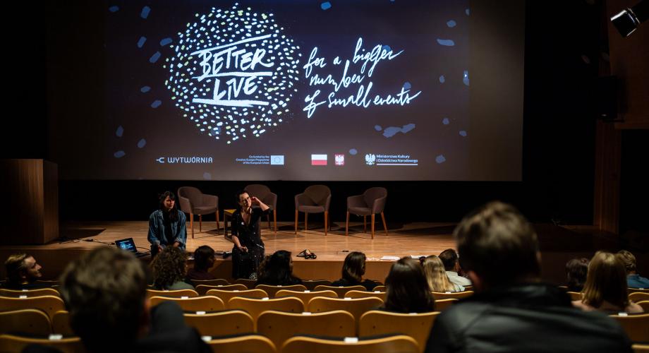 Näkyumä esityssalin takaosasta lavalle, jossa kaksi henkilöä istuu lavan reunalla. Lavalla valkokangas, jossa teksti Better Live for a higher number of small events.