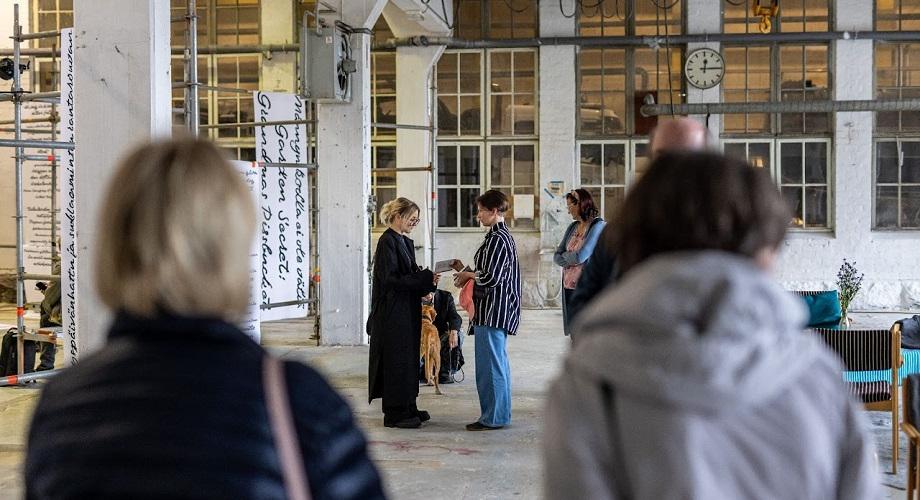 Tapahtuma vanhassa konepajahallissa. Etualalla kaksi osallistujaa selin, kuvan keskellä kaksi henkilöä keskustelevat vastatusten. Taustalla muita osallistujia sekä mustavalkoista tekstiä sisältäviä tekstiplansseja.