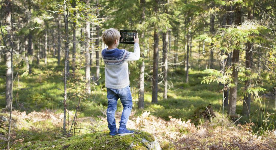 Pojken står med ryggen mot kameran och använder en surfplatta för att fotografera skogen.