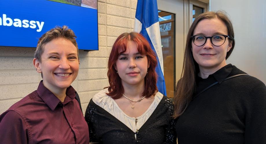 Kolme henkilöä seisoo Suomen lipun ja "embassy"-kyltin edessä.