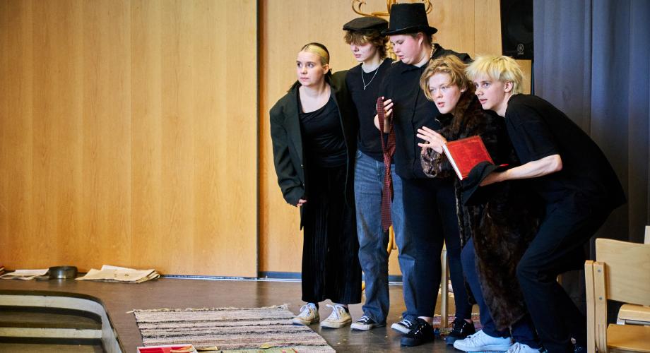 Viisi nuorta seisoo rivissä hieman kyyryssä. Yhdellä on kaulassaan iso puinen risti, toisella kädessään punainen helmikoru ja kolmannella punainen kirja.