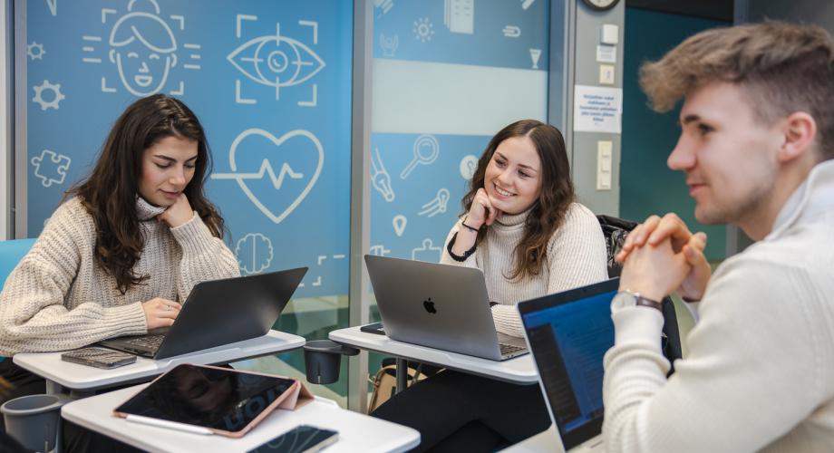 Kuva, jossa kolme hymyilevää nuorta opiskelijaa istuu opetustilassa pöytien ääressä kannettavien tietokoneiden kanssa keskustelemassa ja työskentelemässä yhdessä. Taustalla näkyy sininen seinä, jossa on erilaisia oppimiseen ja teknologiaan liittyviä ikoneita.