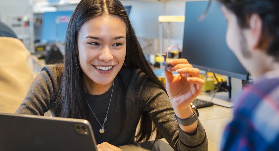 Nainen hymyilee ja pitää kädessään pientä teknistä laitetta. Hän keskustelee toisen henkilön kanssa tietokoneen äärellä, ja taustalla on tekninen laboratorioympäristö.