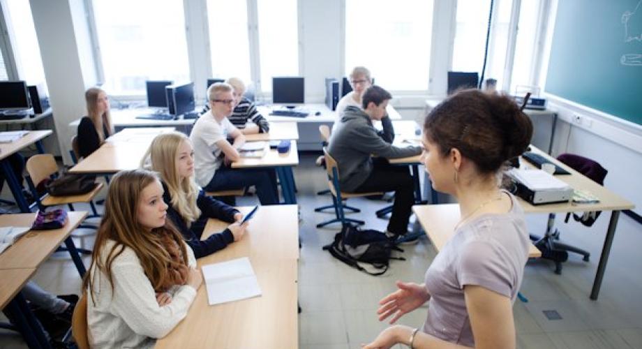 Kuvituskuva koululuokasta, jossa opettaja seisoo ja puhuu luokan etuosassa ja oppilaat kuuntelevat opetusta omilta paikoiltaan pulpettien äärestä.