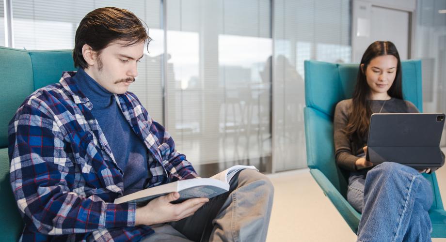 Två studenter sitter i fåtöljer, fördjupade i en bok och en dator.