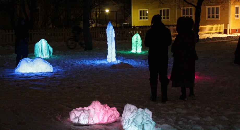 Kaksi henkilöhahmoa pimeässä talvisessa kaupunkimaisemassa, jota valaisee kuudesta erivärisestä ja -muotoisesta valoteoksesta koostuva kokonaisuus.