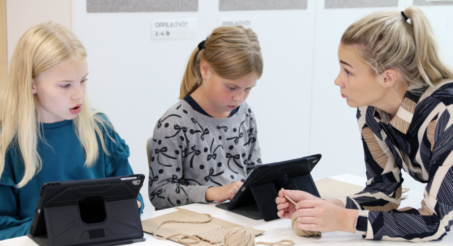 Kuvituskuva, jossa kaksi oppilasta tekee käsityötä opettajan ohjauksessa ja katsoo mallia tabletista.