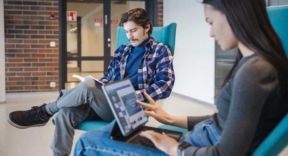 Kuva, jossa kaksi nuorta istuu vierekkäisissä nojatuoleissa sisätiloissa. Toinen lukee kirjaa ja toinen käyttää kannettavaa kannettavaa tietokonetta.