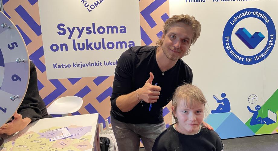 Kuvassa somevaikuttaja Satusetä faninsa Isabella Karvisen kanssa Turun kirjamessujen Lukuloman ja Lukutaito-ohjelman osastolla.
