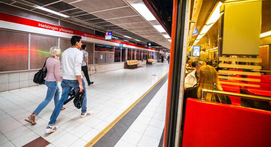 Kävelijöitä metroasemalla ja metron ikkunan läpi näkyviä matkustajia.