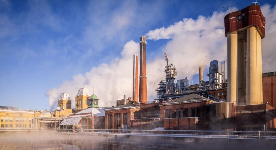 tehdasrakennuksia ja savupiippuja vesialueen takana, ilmassa savua ja höyryä