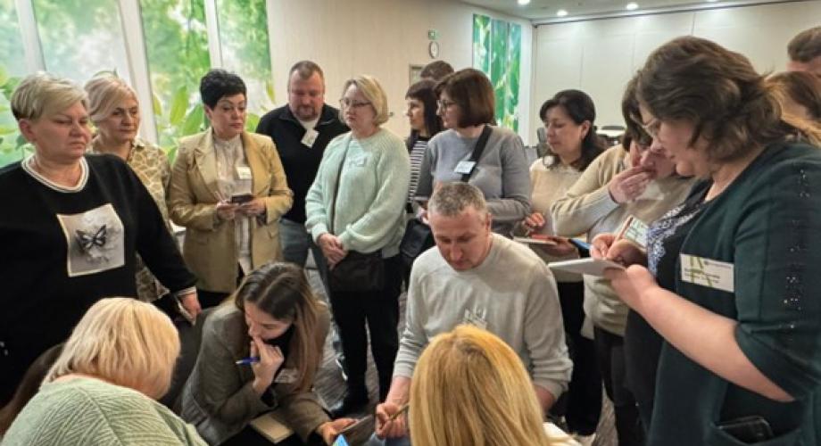 Neljä ammatillisen koulutuksen opettajaa istuu ringissä keskustelemassa aktiivisesti tehtävistä, ja muut opettajat tarkkailevat ryhmätyötä.
