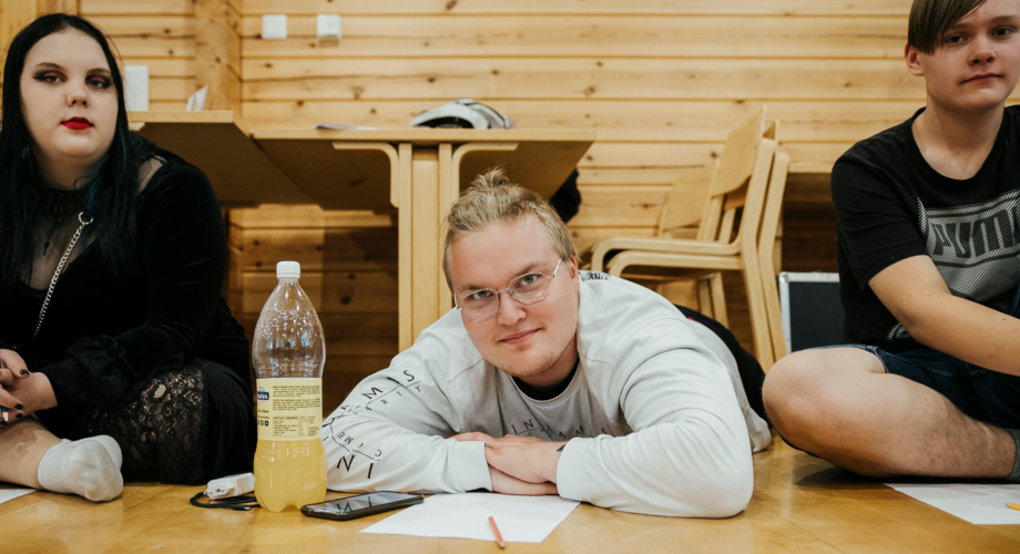Kolme nuorta, joista kaksi istuu ja keskimmäinen loikoilee salin lattialla käsivarsiinsa nojaten.