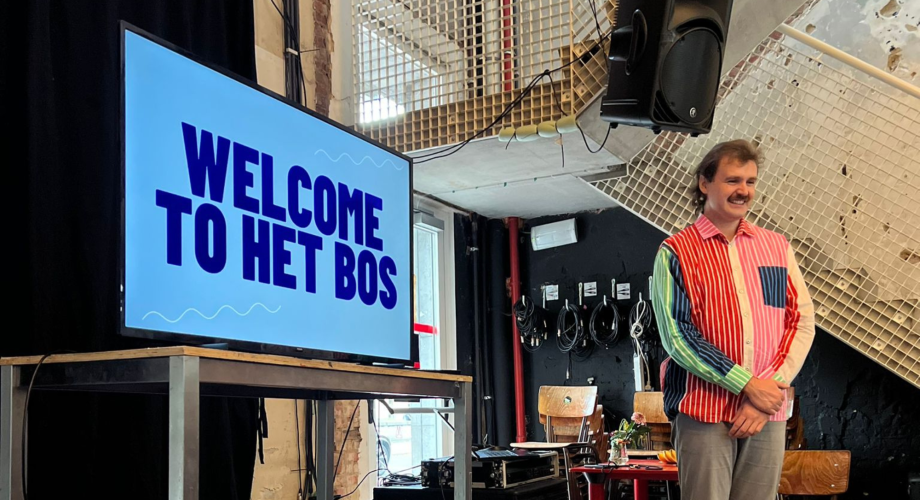 Värikkääseen paitaan pukeutunut mies seisoo näyttöruudun edessä. Näytöllä lukee "Welcome to het bos".