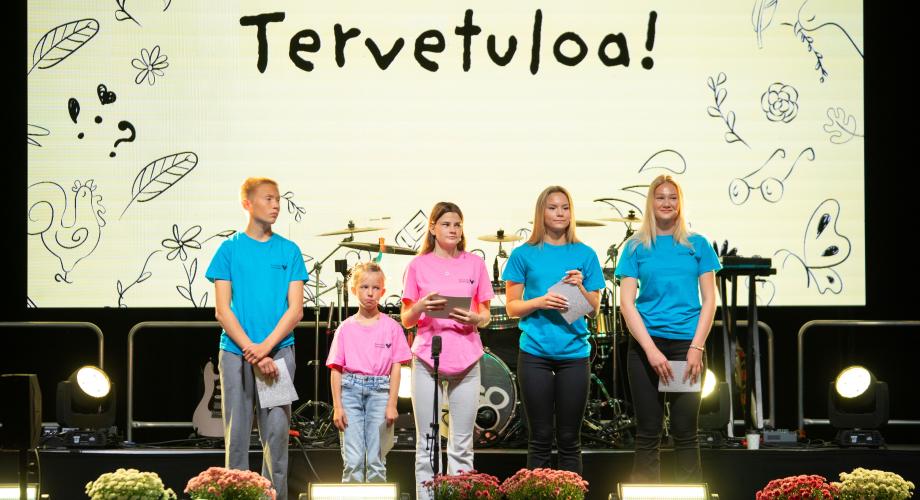 suomeksi: Eri-ikäisiä oppilaita lavalla, taustalla Tervetuloa-kyltti. på svenska: Elever i olika åldrar står på en scen, med en skylt med texten "Tervetuloa" i bakgrunden.