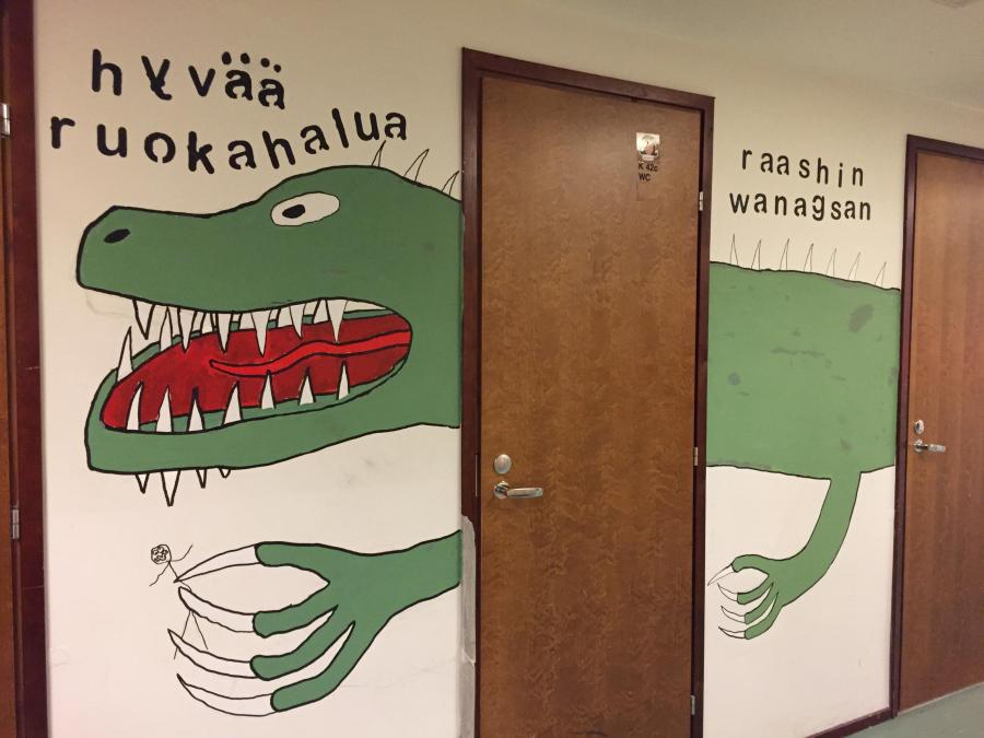 Koulun seinälle maalattu liskohahmo toivottaa hyvää ruokahalua
