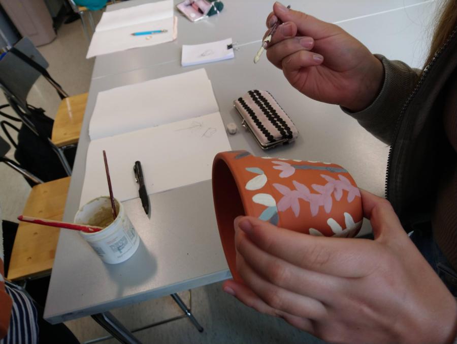Etualalla oikealla oppilas pitelee kädessään saviruukkua, jonka hän on koristellut keramiikkalasitteella kukka-aiheisesti. Toisessa kädessään oppilas pitää koristelussa käyttämäänsä pensseliä ja taustalla näkyy luokkahuone sekä pöytä, jolla on lasitepurkki sekä luonnoslehtiö.