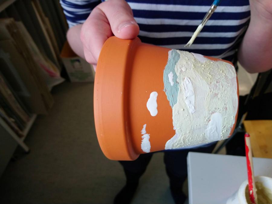 Kuvassa oppilas pitää kädessään saviruukkua, jota hän parhaillaan koristelee maalaten lasitteella ruukun pintaa. Taustalla näkyy luokkahuonetta ja pöydän kulma, jolla on lasitepurkki.