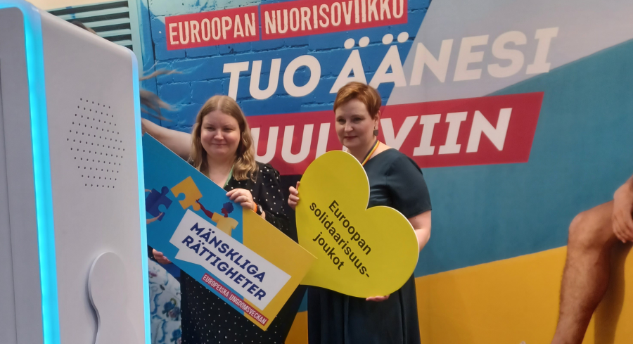 Kaksi henkilöä seisoo messuosastolla kameran edessä ja näyttää kylttejä, joissa lukee "Mänskliga rättigheter" ja "Euroopan solidaarisuusjoukot". Taustalla näkyy värikäs seinä, jossa lukee "Euroopan nuorisoviikko, tuo äänesi kuuluviin"
